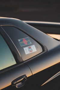 The image captures a close-up view of a car, focusing on the window area. There are stickers visible on the window, which include some text and a graphic of a vehicle. The car is black and the photo is taken at sunset, casting a warm, golden glow on the vehicle's surface.