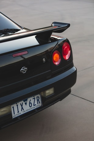 The rear end of a black sports car featuring distinct round taillights and a prominent spoiler is shown. The vehicle is parked on a smooth surface, displaying a license plate that indicates Victoria, Australia.