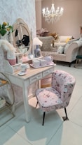 A stylish room with an elegant vanity table and a floral cushioned chair. The table holds decorative items like vases, teacups, and a vintage-style mirror. A chandelier hangs from the ceiling, illuminating the soft, pastel decor. In the background, there is a plush sofa with multiple pillows.