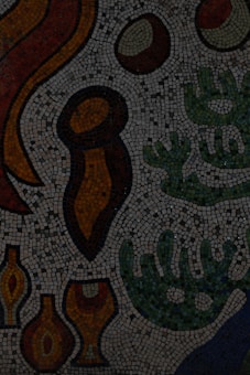 A mosaic artwork composed of various colorful shapes and patterns. The image features abstract forms resembling organic and geometric shapes in shades of red, orange, green, and brown. The intricate design is made up of small, square mosaic tiles against a dark background.