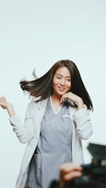 A person with long, dark hair is wearing a light gray professional outfit. They appear joyful, with hair dynamically flowing and a hand raised playfully. The background is light, creating a bright, open atmosphere. Part of a camera or equipment is visible in the foreground, suggesting a photoshoot setting.