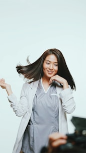 A person with long, dark hair is wearing a light gray professional outfit. They appear joyful, with hair dynamically flowing and a hand raised playfully. The background is light, creating a bright, open atmosphere. Part of a camera or equipment is visible in the foreground, suggesting a photoshoot setting.