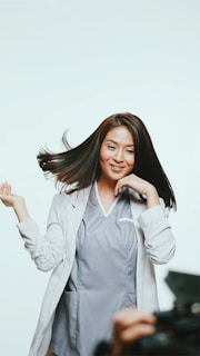 A person with long, dark hair is wearing a light gray professional outfit. They appear joyful, with hair dynamically flowing and a hand raised playfully. The background is light, creating a bright, open atmosphere. Part of a camera or equipment is visible in the foreground, suggesting a photoshoot setting.
