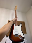 A hand is holding an electric guitar upright in front of a simple background. The guitar has a classic design with a white pickguard and sunburst finish.