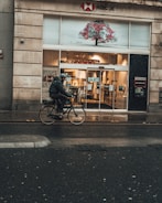 A person wearing a helmet and dark clothing is riding a bicycle on a wet road in front of an HSBC UK bank branch. The building has a large glass entrance with automatic sliding doors. There is a distinctive art piece above the entrance depicting a tree with red elements. The surroundings suggest a city street, and the weather appears to be rainy.