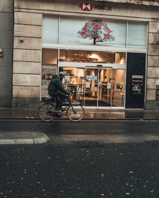 A person wearing a helmet and dark clothing is riding a bicycle on a wet road in front of an HSBC UK bank branch. The building has a large glass entrance with automatic sliding doors. There is a distinctive art piece above the entrance depicting a tree with red elements. The surroundings suggest a city street, and the weather appears to be rainy.