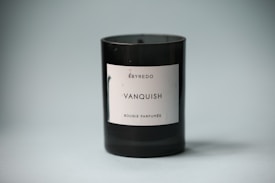 A black cylindrical candle with a white label bearing the text 'BYREDO VANQUISH BOUGIE PARFUMÉE' stands against a soft gray background. The candle's label is clean and minimalistic, giving a modern and sophisticated look.