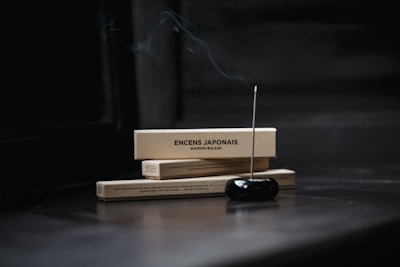 Two rectangular boxes labeled 'ENCENS JAPONAIS' rest horizontally, with a lit incense stick inserted into a black holder in the foreground. Smoke gently rises from the incense, creating a serene and tranquil atmosphere.