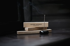 Two rectangular boxes labeled 'ENCENS JAPONAIS' rest horizontally, with a lit incense stick inserted into a black holder in the foreground. Smoke gently rises from the incense, creating a serene and tranquil atmosphere.