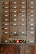 An array of vintage metal mailboxes with numbered labels is arranged in a grid pattern against a wooden backdrop. The mailboxes are slightly weathered, showing signs of age and use. Just below the boxes, a panel features a red cross symbol flanked by two eye-like designs in yellow.