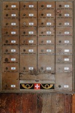 An array of vintage metal mailboxes with numbered labels is arranged in a grid pattern against a wooden backdrop. The mailboxes are slightly weathered, showing signs of age and use. Just below the boxes, a panel features a red cross symbol flanked by two eye-like designs in yellow.