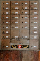 An array of vintage metal mailboxes with numbered labels is arranged in a grid pattern against a wooden backdrop. The mailboxes are slightly weathered, showing signs of age and use. Just below the boxes, a panel features a red cross symbol flanked by two eye-like designs in yellow.
