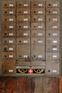 An array of vintage metal mailboxes with numbered labels is arranged in a grid pattern against a wooden backdrop. The mailboxes are slightly weathered, showing signs of age and use. Just below the boxes, a panel features a red cross symbol flanked by two eye-like designs in yellow.