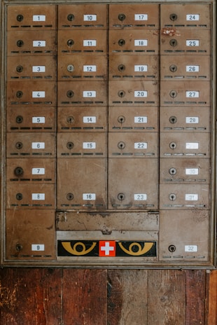 An array of vintage metal mailboxes with numbered labels is arranged in a grid pattern against a wooden backdrop. The mailboxes are slightly weathered, showing signs of age and use. Just below the boxes, a panel features a red cross symbol flanked by two eye-like designs in yellow.