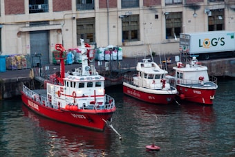 A harbor scene featuring three red fireboats docked side by side along a waterfront. The backdrop consists of an industrial building with bricked upper stories and large windows. To the right, a truck labeled 'LOG'S' is parked, and various bags are stacked against the building.