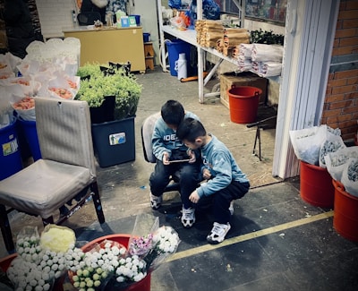 Two children are sitting close together on a chair while focused on a tablet. They appear to be in a flower shop or market setting, as various flower bouquets in buckets, along with other items like paper-wrapped bundles, are visible around them. A variety of colors can be seen in the flowers, including whites, greens, and pinks. The environment appears somewhat cluttered and industrial.