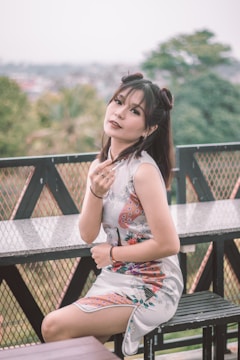 A person is seated outdoors on a bench wearing a floral-patterned white dress. Fencing and green foliage can be seen in the background, suggesting a garden or park setting. The person's hair is styled in an updo with loose strands.