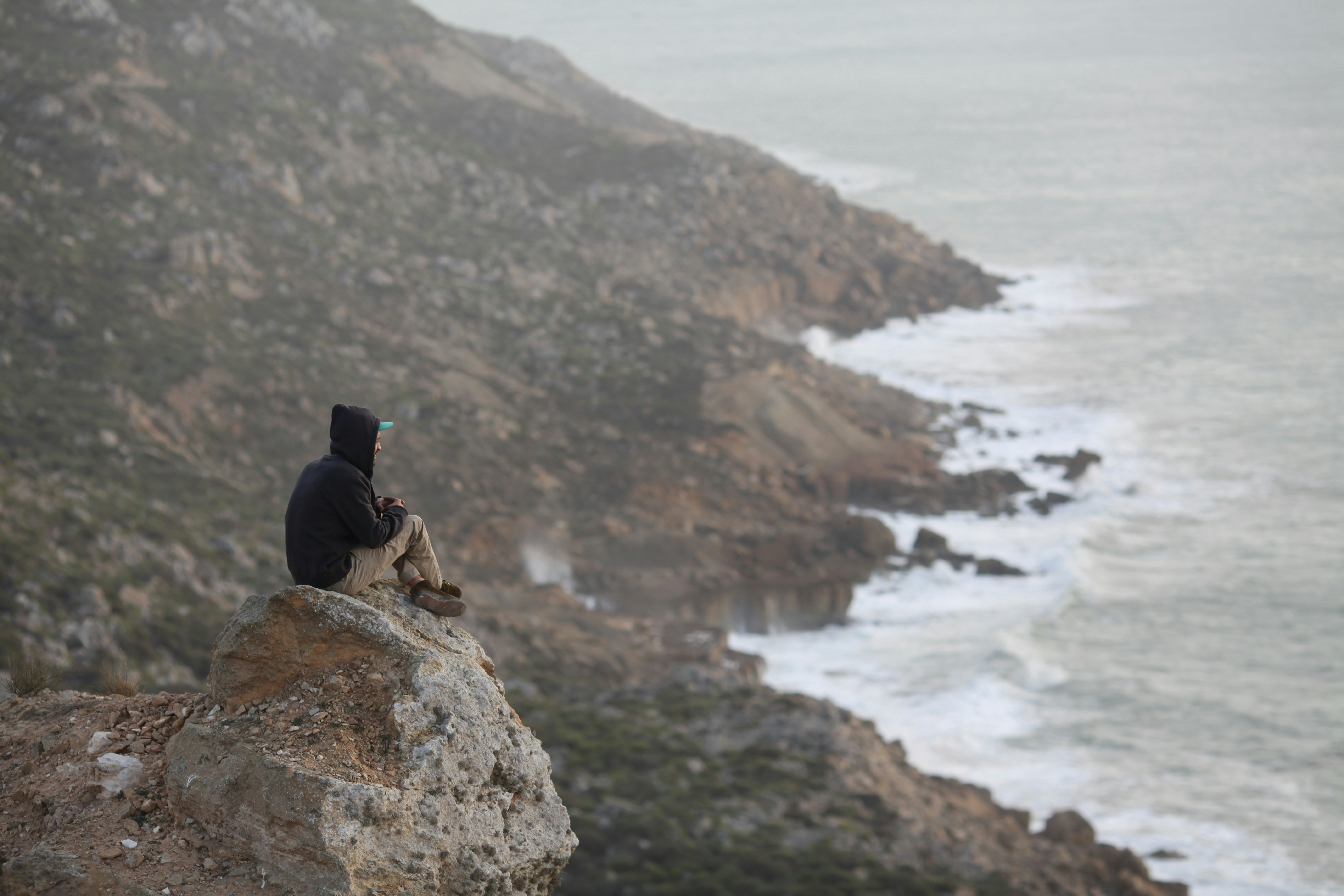 man in black jacket sitting on rock formation near body of water during daytime