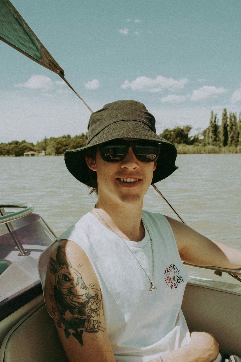 Mann in weißem Tanktop mit schwarzer Sonnenbrille und braunem Hut sitzt tagsüber auf dem Boot