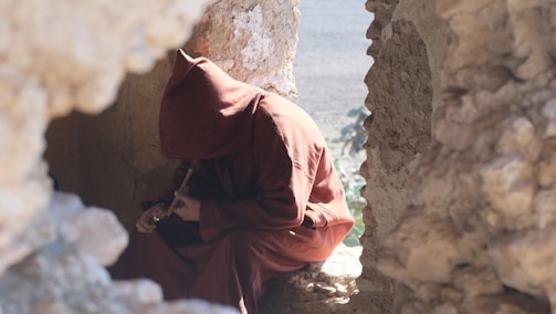 A person wearing a hooded brown robe is sitting in a rocky, ancient stone structure. The individual is holding a small flute. The scene is lit by natural light, giving a contemplative and mysterious atmosphere, as the person appears to be engaged in a solitary moment.