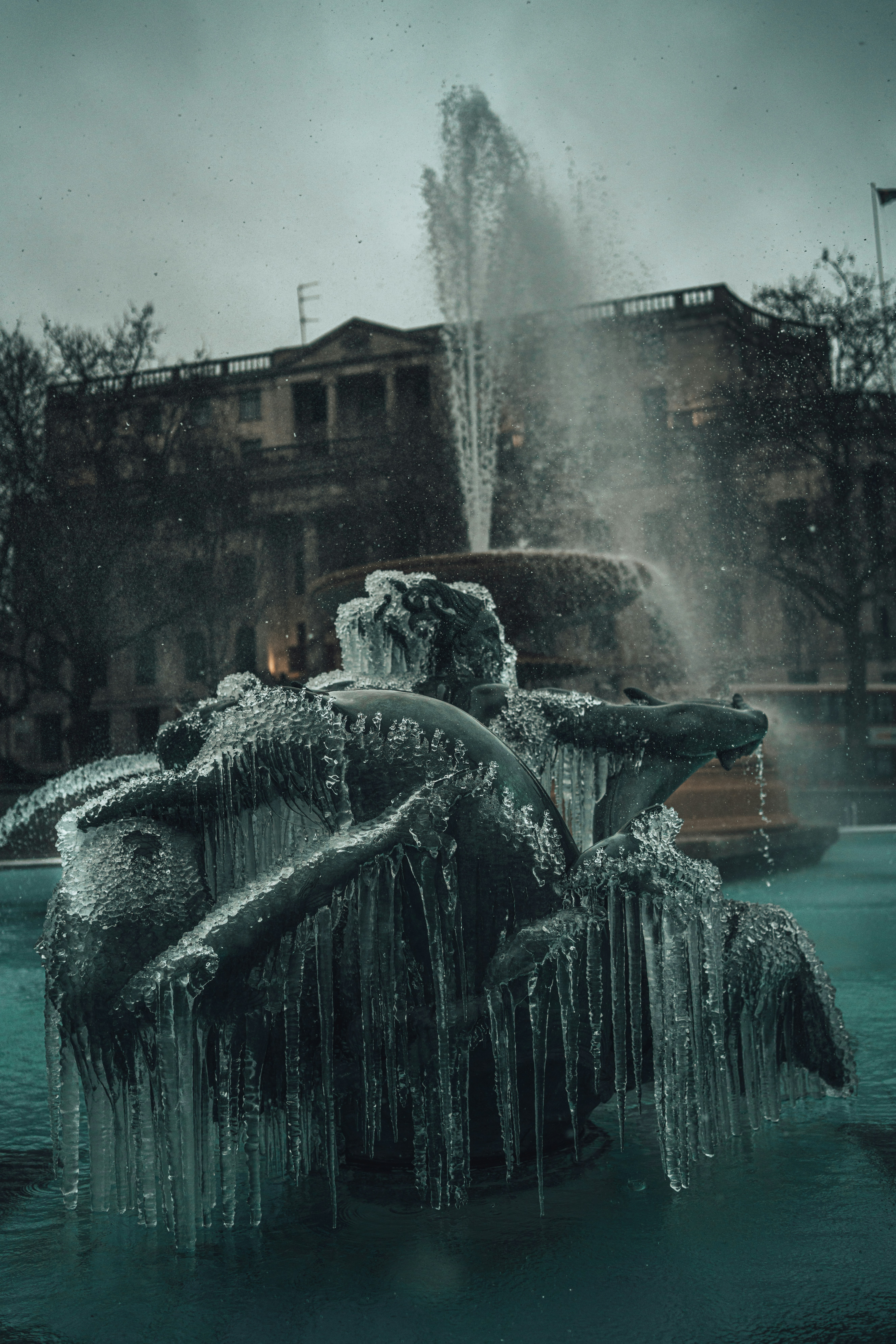 A fountain slowly thawing, with water flowing beneath sheets of ice in front of a grand old building—frozen and alive at the same time.