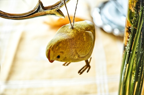 A small wooden or ceramic bird ornament is suspended by a string, hanging from a decorative branch. The background is softly blurred, showing hints of a table with a cloth and a vase containing stems.