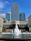 A large fountain with multiple water jets surrounded by a stone basin is in the foreground. Behind the fountain, there are modern skyscrapers, with one prominently visible in the center, flanked by buildings with HSBC and Citi logos. The sky is clear and blue, and a few people are sitting or walking around the area.