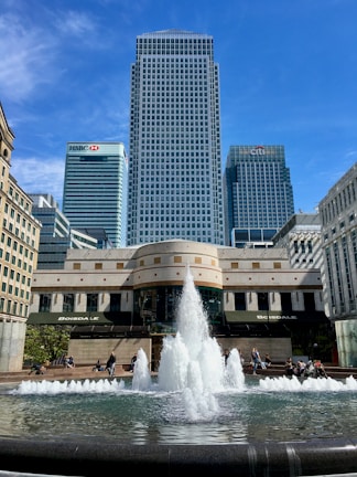 A large fountain with multiple water jets surrounded by a stone basin is in the foreground. Behind the fountain, there are modern skyscrapers, with one prominently visible in the center, flanked by buildings with HSBC and Citi logos. The sky is clear and blue, and a few people are sitting or walking around the area.