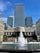 A large fountain with multiple water jets surrounded by a stone basin is in the foreground. Behind the fountain, there are modern skyscrapers, with one prominently visible in the center, flanked by buildings with HSBC and Citi logos. The sky is clear and blue, and a few people are sitting or walking around the area.