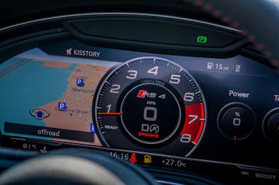 A digital car dashboard displays a map with navigation, speedometer, and various gauges. The map shows detailed routes and landmarks. The central tachometer shows RPM with a redline indication and branding. Additional information includes mileage, fuel level, power meter, and temperature.