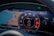 A digital car dashboard displays a map with navigation, speedometer, and various gauges. The map shows detailed routes and landmarks. The central tachometer shows RPM with a redline indication and branding. Additional information includes mileage, fuel level, power meter, and temperature.