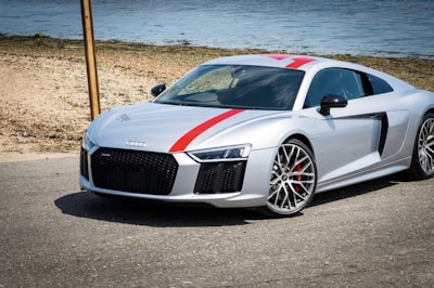 A sleek silver sports car is parked on an asphalt road along a beach setting. It features distinctive red racing stripes and large, stylish alloy wheels. The backdrop includes a tranquil body of water and a sandy shoreline.