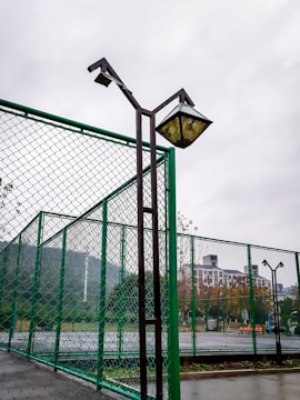 A fenced sports court with high metal mesh barriers in the foreground and background. Tall lampposts stand adjacent to the fence, showing wear and discoloration. Beyond the fence, there are trees with autumn foliage and a high-rise building partially visible through overcast and cloudy weather.
