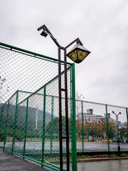 A fenced sports court with high metal mesh barriers in the foreground and background. Tall lampposts stand adjacent to the fence, showing wear and discoloration. Beyond the fence, there are trees with autumn foliage and a high-rise building partially visible through overcast and cloudy weather.