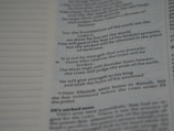 An open page with printed text from a religious book, possibly a Bible, featuring distinct verses and organized lines. The text is black on a white background.