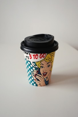 A disposable coffee cup with a pop art illustration of a woman holding a phone, featuring vibrant colors and comic-style design. The text '5 TO GO' is prominently displayed.