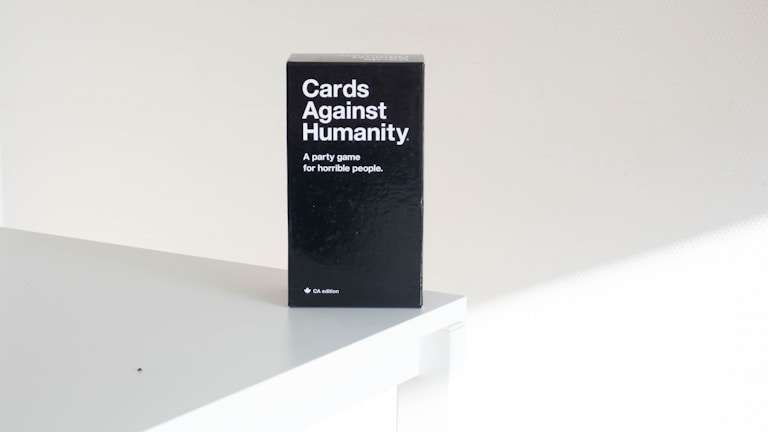 A black box of the card game 'Cards Against Humanity' is placed on a white surface. The text on the box reads 'A party game for horrible people'. The setting appears minimalistic with a focus on the game's packaging.