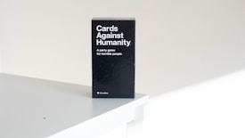 A black box of the card game 'Cards Against Humanity' is placed on a white surface. The text on the box reads 'A party game for horrible people'. The setting appears minimalistic with a focus on the game's packaging.