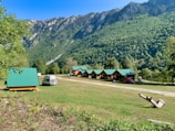 A serene landscape featuring small wooden cabins with green roofs situated on a grassy field. In the foreground, a white camper van is parked alongside a few people. The background is dominated by lush green mountains under a clear blue sky. A piece of driftwood lies on the ground to the right.
