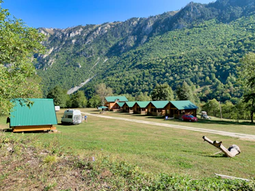 A serene landscape featuring small wooden cabins with green roofs situated on a grassy field. In the foreground, a white camper van is parked alongside a few people. The background is dominated by lush green mountains under a clear blue sky. A piece of driftwood lies on the ground to the right.
