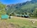 A serene landscape featuring small wooden cabins with green roofs situated on a grassy field. In the foreground, a white camper van is parked alongside a few people. The background is dominated by lush green mountains under a clear blue sky. A piece of driftwood lies on the ground to the right.