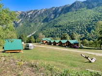 A serene landscape featuring small wooden cabins with green roofs situated on a grassy field. In the foreground, a white camper van is parked alongside a few people. The background is dominated by lush green mountains under a clear blue sky. A piece of driftwood lies on the ground to the right.