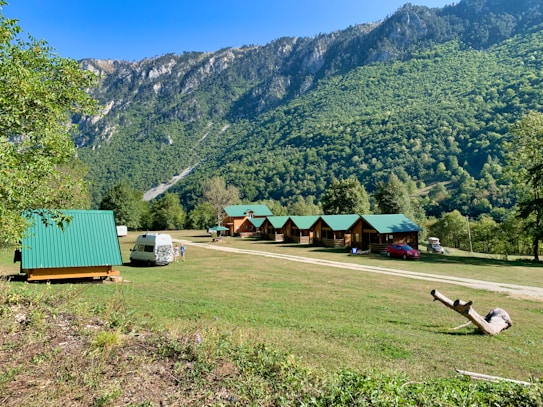 A serene landscape featuring small wooden cabins with green roofs situated on a grassy field. In the foreground, a white camper van is parked alongside a few people. The background is dominated by lush green mountains under a clear blue sky. A piece of driftwood lies on the ground to the right.