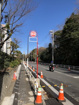 A road with traffic cones lined up along the sides and a red signpost in the center. The road is bordered by trees and vegetation on one side and a sidewalk on the other. A person on a motorbike is driving on the road. There are power lines running overhead, and the sky is clear and blue.