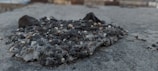 A small mound of rough, uneven pebbles and gravel sits atop a concrete surface. The pebbles vary in color and size, ranging from small white and beige stones to larger dark gray rocks. The surface appears weathered and slightly uneven, capturing a raw and natural texture.
