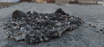 A small mound of rough, uneven pebbles and gravel sits atop a concrete surface. The pebbles vary in color and size, ranging from small white and beige stones to larger dark gray rocks. The surface appears weathered and slightly uneven, capturing a raw and natural texture.