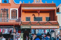 A vibrant street scene features a colorful building with terracotta roofing. The facade has decorative windows and signage reading 'Most Complete Store' and 'Mexico Curios.' An Iron Man figurine is attached to an upper balcony. Below, clothing and souvenirs are displayed outside the store, with people browsing.
