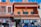 A vibrant street scene features a colorful building with terracotta roofing. The facade has decorative windows and signage reading 'Most Complete Store' and 'Mexico Curios.' An Iron Man figurine is attached to an upper balcony. Below, clothing and souvenirs are displayed outside the store, with people browsing.