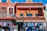 A vibrant street scene features a colorful building with terracotta roofing. The facade has decorative windows and signage reading 'Most Complete Store' and 'Mexico Curios.' An Iron Man figurine is attached to an upper balcony. Below, clothing and souvenirs are displayed outside the store, with people browsing.