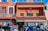 A vibrant street scene features a colorful building with terracotta roofing. The facade has decorative windows and signage reading 'Most Complete Store' and 'Mexico Curios.' An Iron Man figurine is attached to an upper balcony. Below, clothing and souvenirs are displayed outside the store, with people browsing.